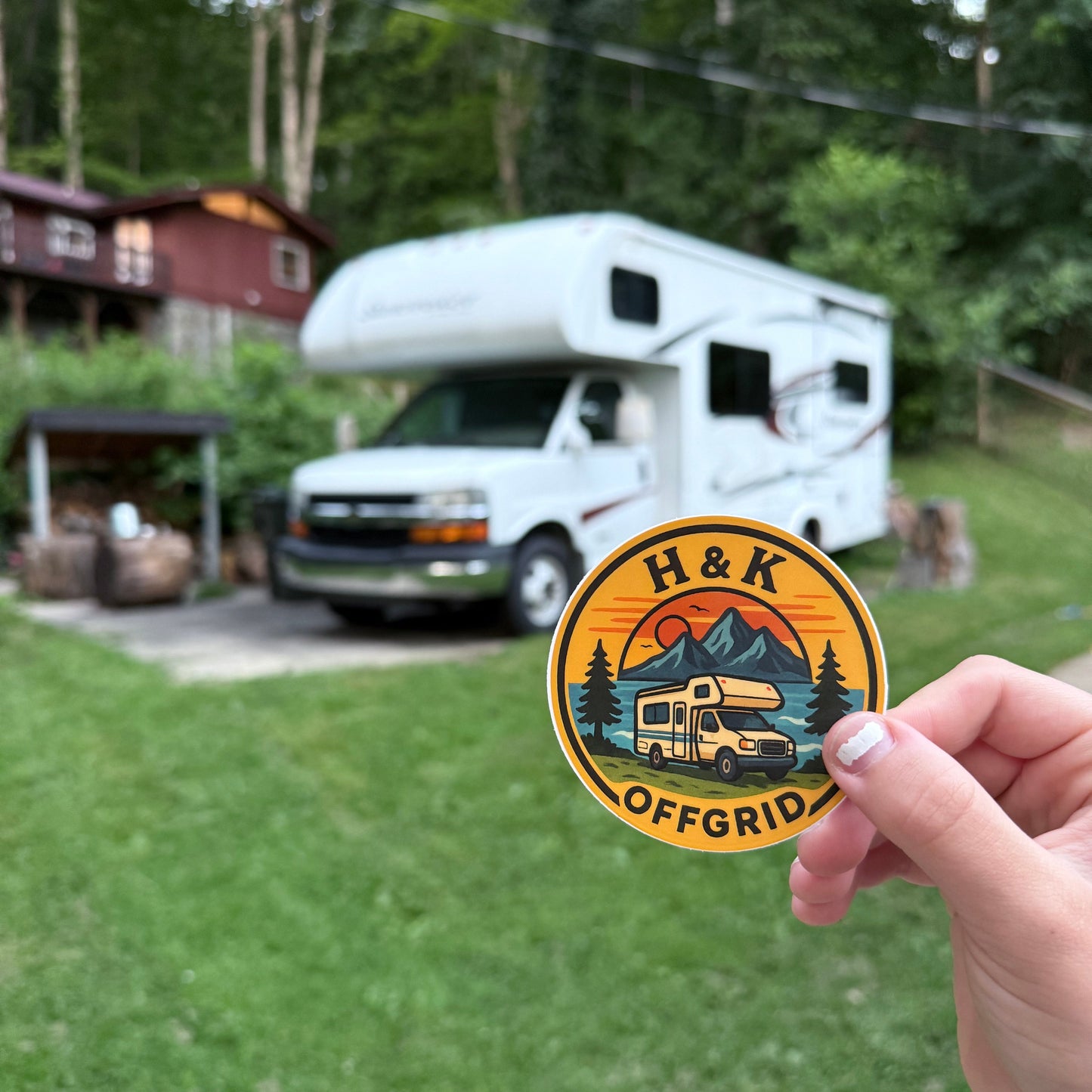 Hand holding a round sticker with 'H & K OFFGRID' logo in front of an RV.