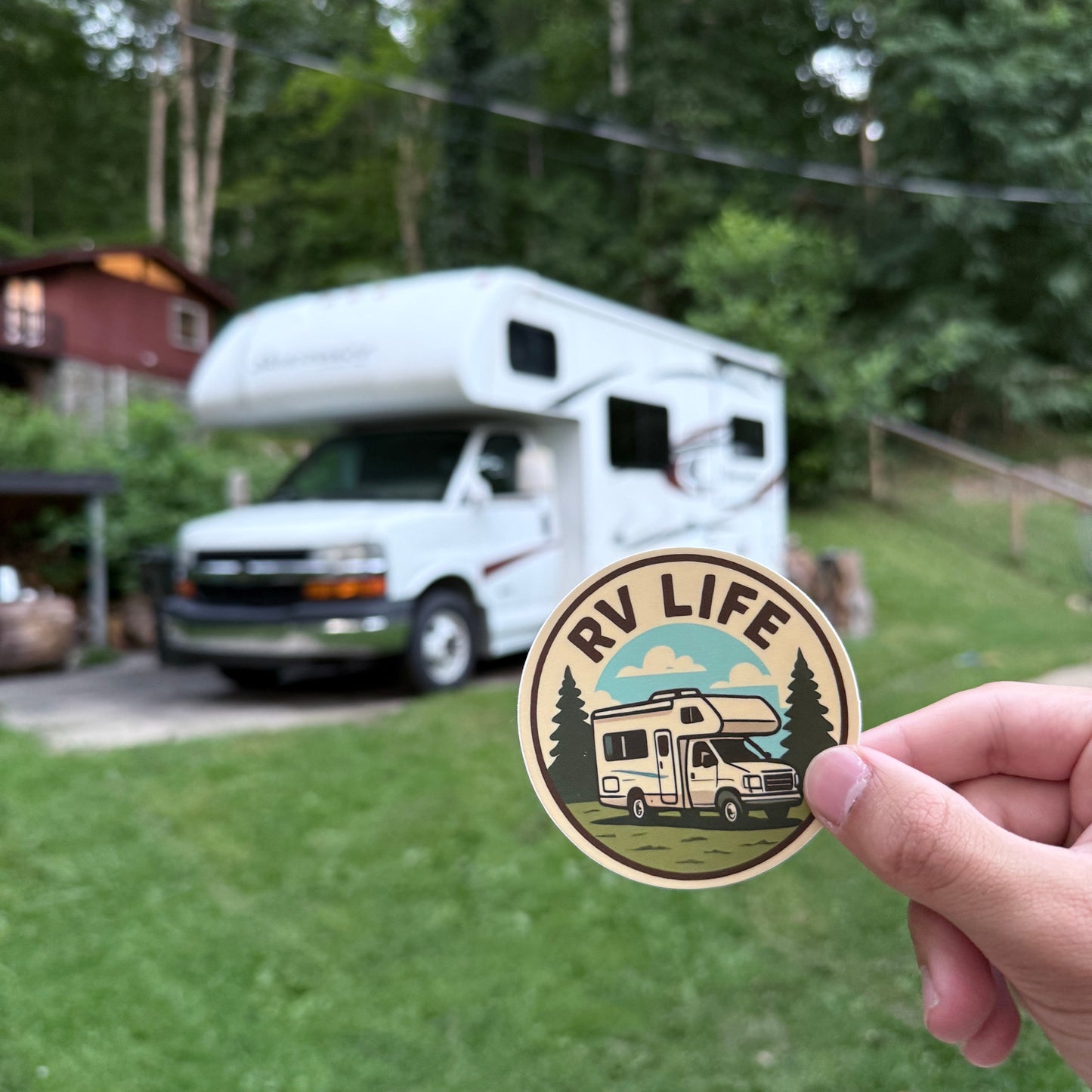 Hand holding a round sticker with 'RV Life' design in front of a white RV parked outdoors.